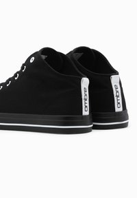 Pár černých plátěných tenisek s bílými očky, bílými patkami s nápisem "ombre" a černými podrážkami s bílými pruhy, zobrazené zezadu.