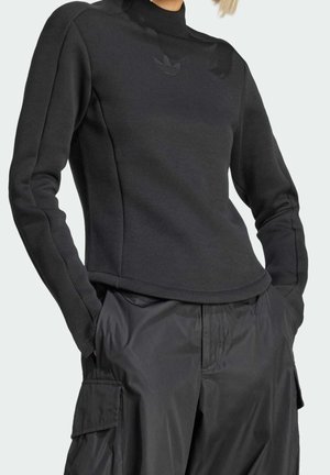 Femme portant un haut noir ajusté à manches longues et un pantalon cargo noir ample, les mains dans les poches, sur un fond uni.