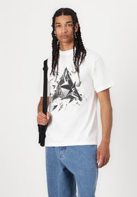 White cotton t-shirt with a graphic black and metallic print in the shape of a fractal triangle, paired with blue denim jeans and a black strap bag.