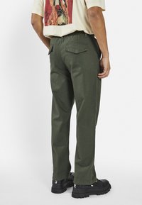 Olive green pants with a straight cut, textured fabric, and two back pockets. Worn with black footwear, seen from the back.