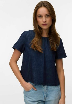 Jeune femme aux longs cheveux bruns portant un haut bleu marine à manches courtes et un jean bleu clair, une main dans la poche.