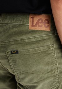 Close-up of a person wearing olive green corduroy pants with a visible Lee brand leather patch and stitched back pocket.