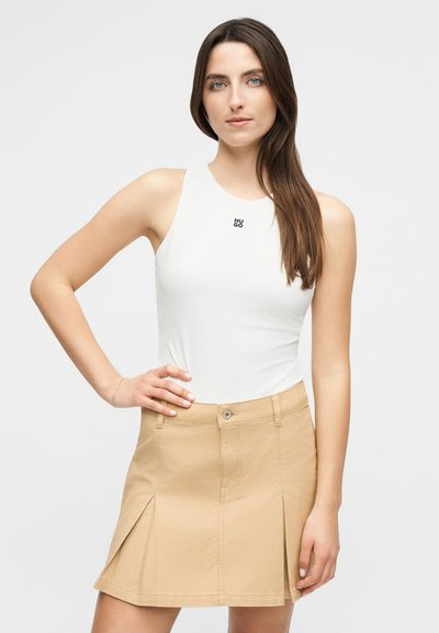 Femme aux longs cheveux bruns portant un top blanc sans manches avec le logo "HUGO" et une jupe plissée beige, debout avec une main sur la hanche.