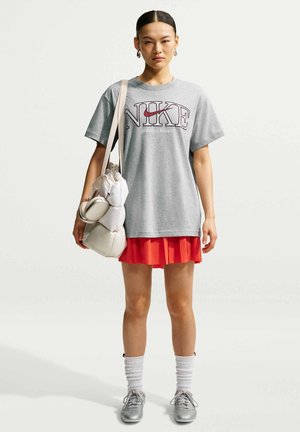Vrouw met zwart haar in een paardenstaart draagt een grijs oversized Nike T-shirt, een rode geplooide rok, witte sokken, zilveren sneakers en draagt een beige tas.