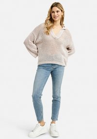 Hellrosa gestrickter Pullover mit lockerer Passform, V-Ausschnitt und offener Maschenstruktur, kombiniert mit hellblauen Jeans und weißen Sneakern.