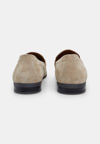 Beige Wildlederschuhe mit einem niedrigen schwarzen Absatz, die eine glatte Textur und eine abgerundete Ferse aufweisen. Sie sind schlicht gestaltet, ohne sichtbare Verzierungen oder Muster.
