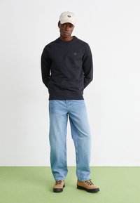 Felpa nera con scollo rotondo, jeans in denim blu leggeri e stivali beige con lacci. Cappellino beige con dettagli del logo.