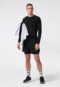 Mann in schwarzer Sportbekleidung hält einen grauen Sweatshirt über der Schulter, trägt schwarze Shorts, schwarze Sneakers und weiß gestreifte Socken und steht vor einem weißen Hintergrund.