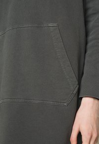 Närbild på en mörkgrå huvtröjeficka med synlig sömnad bredvid en avslappnad hand och uppvikt ärm.