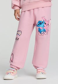 Pantaloni della tuta rosa in morbido tessuto, decorati con personaggi dei cartoni animati sulla gamba, grafiche colorate e polsini a costine con un dettaglio a forma di cuore.