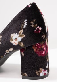 Bloc-chaussure en velours noir avec motif floral texturé en tons blanc, rouge et marron, sur fond blanc uni.