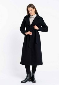 Długi czarny wełniany płaszcz o dwurzędowym kroju, szerokich klapach i dopasowanym pasku, zestawiony z czarnymi wąskimi spodniami i botkami za kostkę.