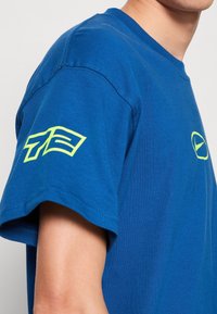 Modré bavlněné tričko s krátkými rukávy. Na levém rukávu se nachází zelená grafika a na hrudi je menší logo. Hladký povrch.