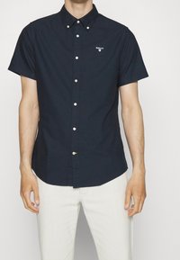 Granatowa koszula z krótkim rękawem na guziki, wykonana z bawełny, z kołnierzykiem o spiczastym kształcie, białym logo na piersi i prostym dołem.