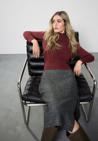 Juoda odinė kėdė su chromuota konstrukcija; modelis dėvi bordo spalvos golfą ir pilką austą sijoną; glotnūs plaukai; minkštas apšvietimas ir minimalistinė fonas.