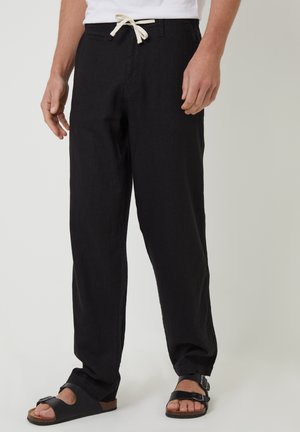 Homme portant un pantalon noir ample avec un cordon blanc et des sandales noires à bouts ouverts sur un fond clair.