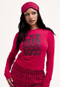 Piros hosszú ujjú felső vastag fekete betűkkel: "I’VE BEEN SOOO GOOD." A hozzáillő piros nadrágon fekete szív- és csillagminták találhatók. Tartozik hozzá egy koordinált fejpánt is.