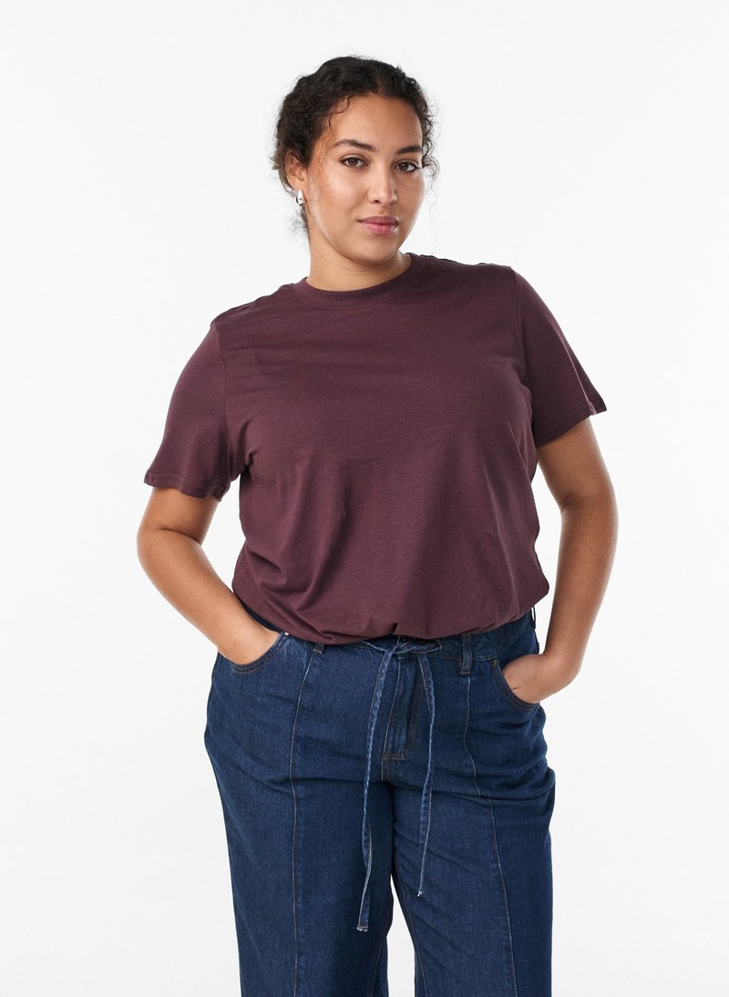 Femme aux cheveux bouclés portant un t-shirt à manches courtes bordeaux et un pantalon en jean bleu taille haute, les mains dans les poches, debout devant un fond blanc.