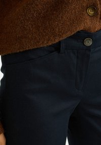 Pantalones azul oscuro de tela suave, con corte ajustado, bolsillos laterales y un botón decorativo dorado con diseño geométrico.