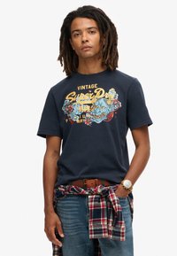 Camiseta de algodón azul marino con un diseño gráfico vintage en amarillo y azul. El modelo lleva una camisa de cuadros atada a la cintura.
