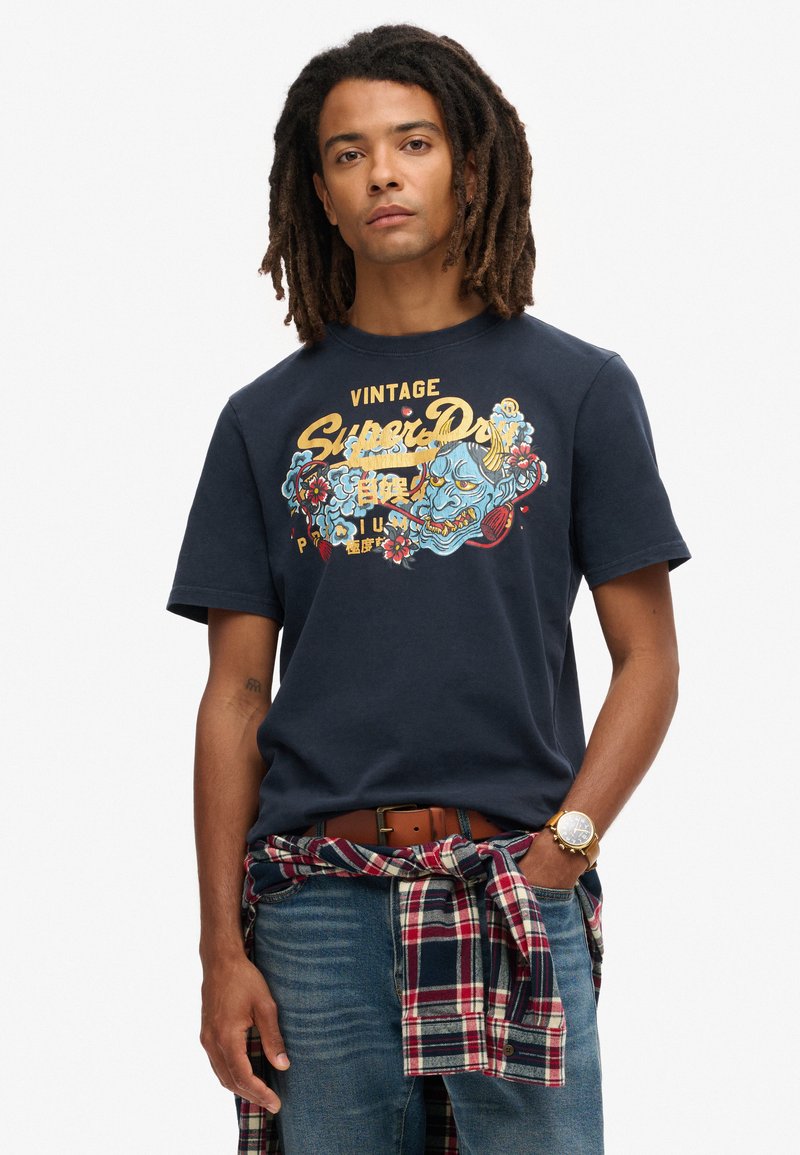 Camiseta de algodón azul marino con un diseño gráfico vintage en amarillo y azul. El modelo lleva una camisa de cuadros atada a la cintura.