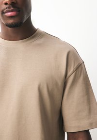 Beež lühikeste varrukatega T-särk, mis on valmistatud pehmest puuvillast, ümar kaelusega ja lõdva lõikega, õlal peente õmbluste detailidega.