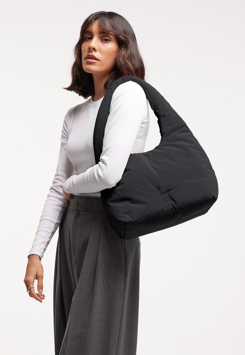 Vrouw met schouderlang donker haar die een wit shirt met lange mouwen en grijze broek draagt, met een grote zwarte gewatteerde schoudertas.