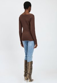 Brauner gestrickter Langarmoberteil kombiniert mit hellblauen Skinny-Jeans und kniehohen Schlangenprint-Absatzstiefeln, von hinten betrachtet.