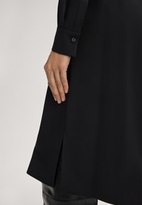 Zwarte dress van een soepel materiaal, met lange mouwen en een knoopmanchet. Zijsplitten verbeteren de bewegingsvrijheid. Gladde textuur met minimale accenten.