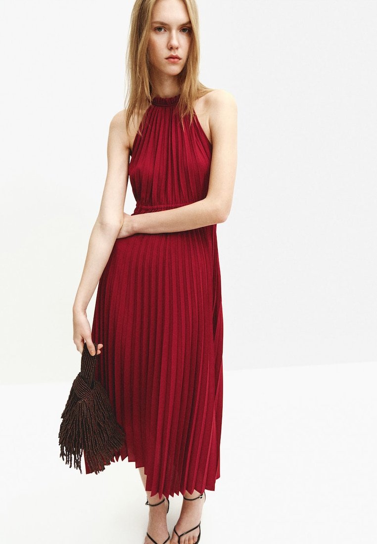 Jeune femme en robe midi rouge plissée sans manches, tenant un sac à main marron foncé texturé, portant des sandales noires à brides, sur fond blanc uni.
