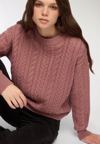 Sweter w warkoczowym splocie w kolorze przybrudzonego różu, z ściągaczami na mankietach i dekolcie. Miękka faktura z wyraźnymi wzorami; noszony na czarnych spodniach.