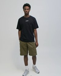 Młody mężczyzna stojący, ubrany w czarną koszulkę z logo, oliwkowe krótkie spodnie typu cargo, białe skarpetki i srebrne sneakersy na jednolitym jasnym tle.