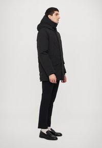 Parka neagră căptușită cu guler înalt și glugă, având închidere cu fermoar pe față și buzunare laterale, purtată peste pantaloni negri mulați și încălțări.