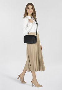 Borsa a tracolla nera con superficie texturizzata, chiusura con zip e tracolla regolabile, abbinata a una blusa bianca e una gonna plissettata beige.