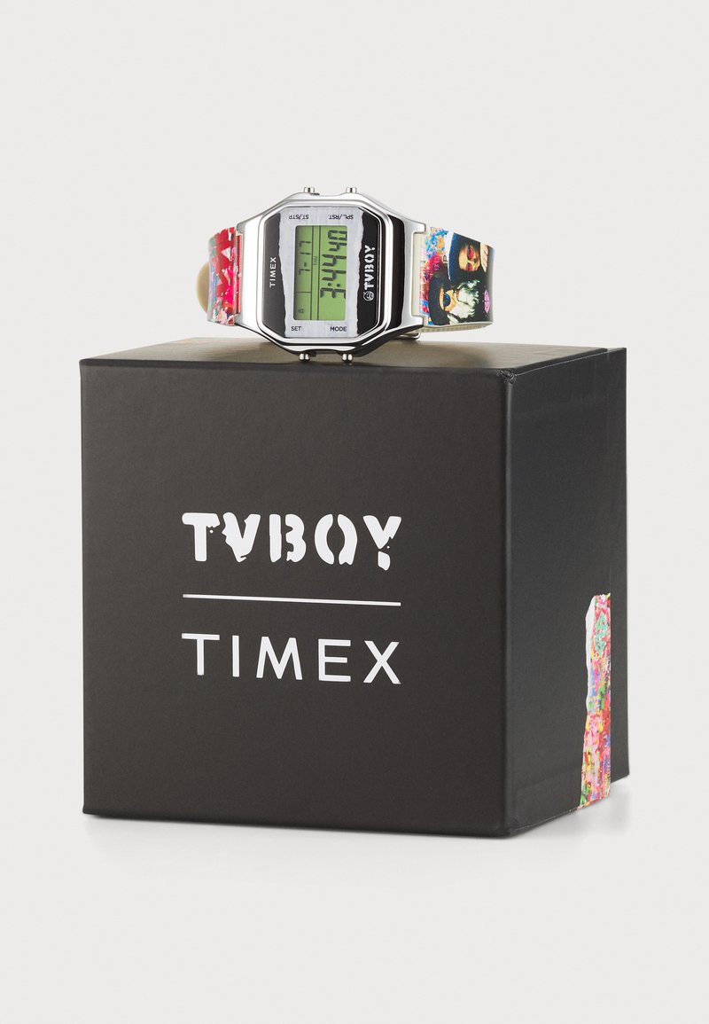 Reloj digital con caja de plata, pantalla verde y correa con patrón colorido. Colocado sobre una caja negra con los logotipos de "TVBOY" y "TIMEX".