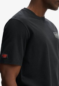 Camiseta de algodón negra con cuello redondo, mangas cortas y un parche en la manga izquierda con la marca "SUPERDRY" en rojo sobre un fondo negro.
