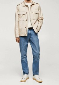 Beżowa bawełniana kurtka z dwoma kieszeniami na piersi, brązowymi guzikami i naramiennikami, zestawiona z niebieskimi dżinsami i białymi trampkami.