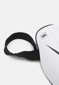 Hvid plastik skinnebeskytter med et sort Nike-logo og "NIKE CHARGE"-mærkat. Udstyret med en sort Velcro-rem for sikker fastgørelse. Glat overflade.