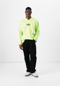Dolga majica z dolgimi rokavi v neonsko zeleni barvi z V-izrezom, z osrednjim črnim logotipom Adidas na prsih, v kombinaciji s črnimi hlačami in svetlimi športnimi copati.
