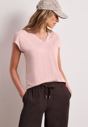Donna con top rosa chiaro a maniche corte e scollo a V, pantaloni marrone scuro con coulisse e cappellino beige con fantasia, posa con le mani nelle tasche.