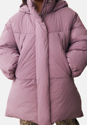 Femme portant un manteau d'hiver matelassé rose poudré avec les mains dans les poches et un pantalon imprimé léopard, debout devant un fond uni.