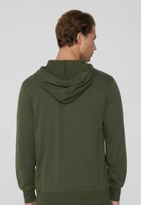 Sudadera verde oscura hecha de una tela suave, con un corte relajado, capucha y mangas largas. La parte trasera presenta un diseño sencillo sin patrones.