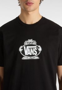 Černé bavlněné tričko s velkým bílým logem Vans a kreslenou postavičkou, která drží ovál. Kulatý výstřih a krátké rukávy.