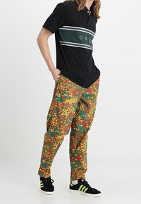 Svart polo-shirt med gröna och vita ränder, parat med löst sittande cargobyxor i en flerfärgad prickig mönster, bärs med svarta sneakers.