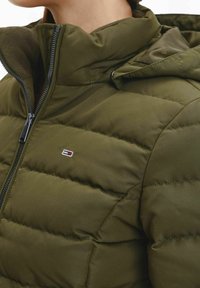 Olivgrön vadderad jacka med hög krage, med horisontella quiltade sektioner och dragkedja framtill. Liten logga på bröstet.