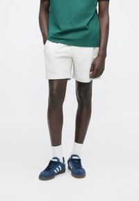 Calções brancos de algodão, t-shirt verde, ténis de camurça azul com riscas azul claro, meias brancas. Design casual simples, corte ajustado.