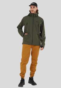 Olijfgroene waterdichte jas met een rits aan de voorkant, een verstelbare capuchon en zwarte accenten. Gecombineerd met oranje broek en zwarte schoenen.