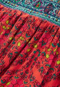 Gros plan sur un tissu orange avec un motif floral vert, rose et jaune et une bordure brodée bleu et violet délicate.