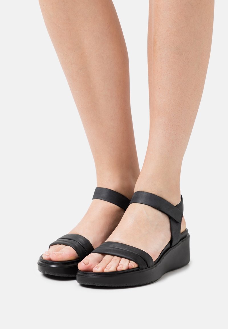 Pieds portant des sandales compensées noires avec des brides à la cheville et des orteils ouverts sur un fond clair.