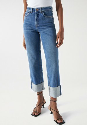 Frau trägt hoch taillierte blaue Jeans mit breiten umgeschlagenen Säumen und schwarzen, mit Riemchen versehenen Sandaletten mit Absatz, steht vor einem weißen Hintergrund.